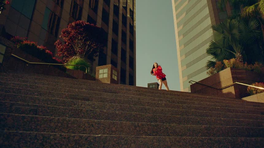 A woman is outside in front of buildings as she moves her arms then pulls her foot back to kick