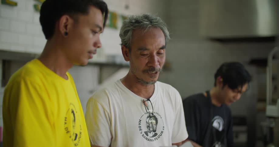 A young man stirs flour in a bowl while speaking to the older man standing beside him in an industrial kitchen