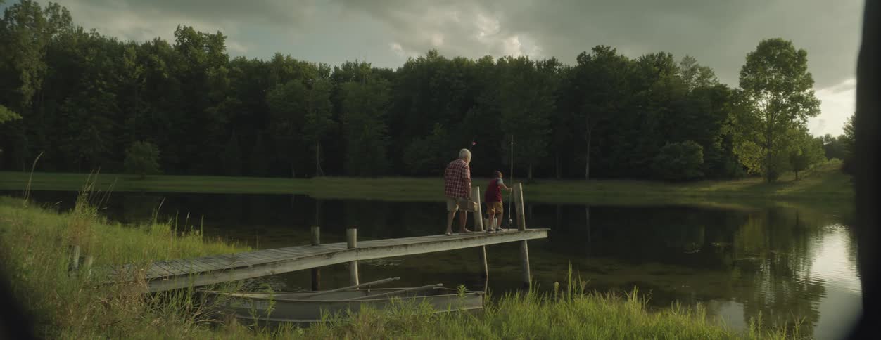 A graying man and a boy walk along a pier towards a lake while carrying fishing equipment