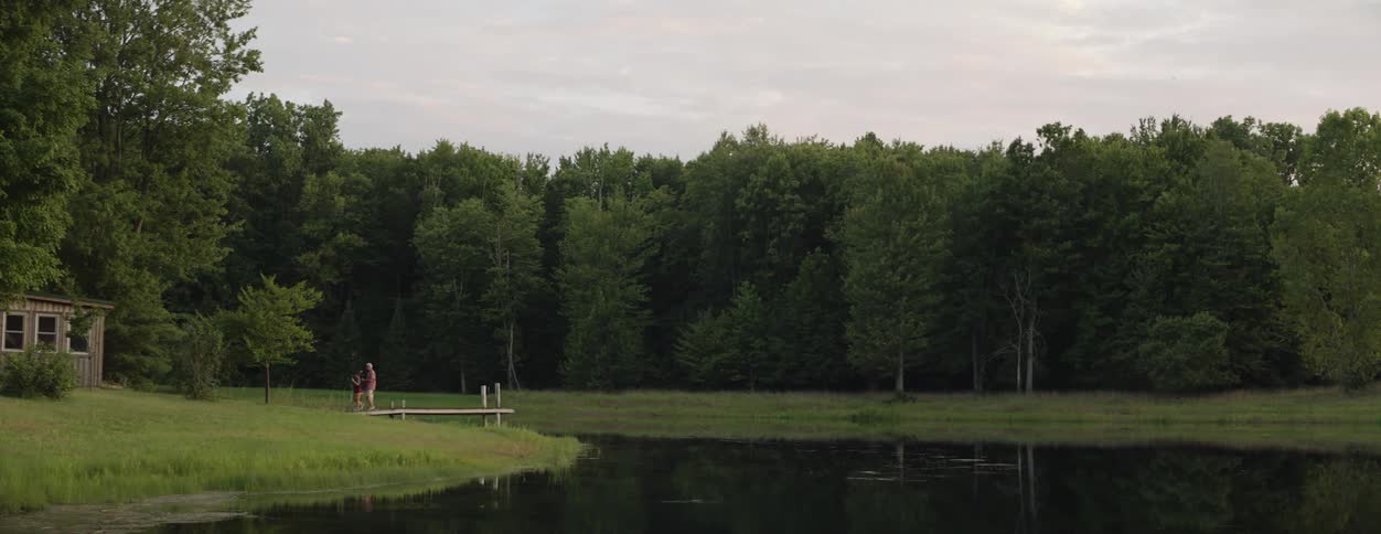 A young boy and a man are standing on a dock overlooking a pond and they walk to a nearby cabin surrounded by woods