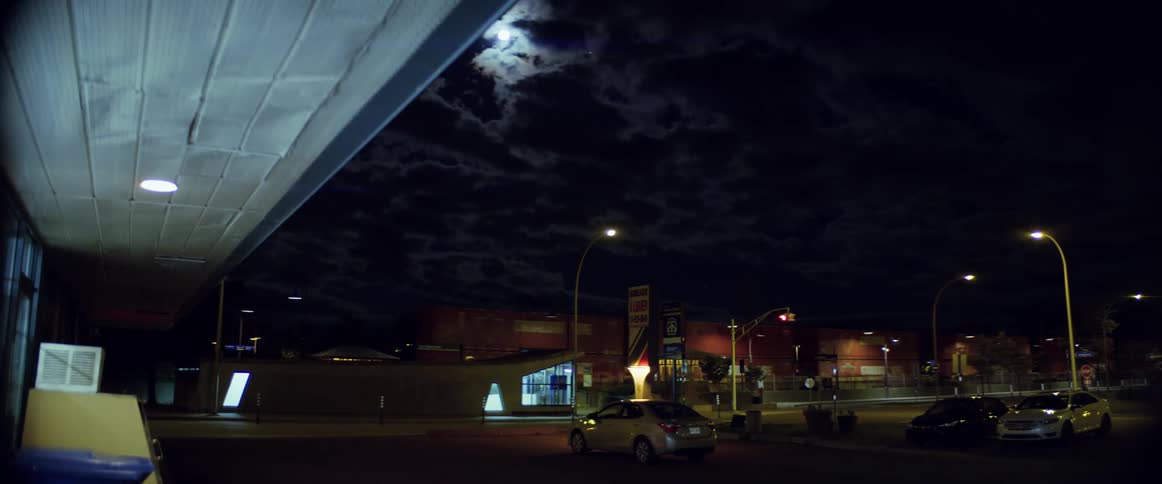 A cargo train travels past stores and a parking lot at night