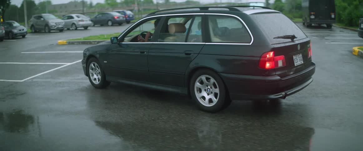 An automobile with two people inside travels across a wet parking lot as the teenager in the back seat drinks from a straw in a cup