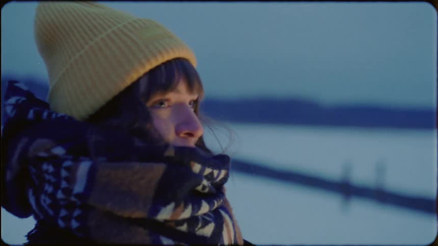 A woman is standing outside in the snow wearing a coat and hat with a scarf wrapped around her face and she is rubbing her arms trying to warm herself