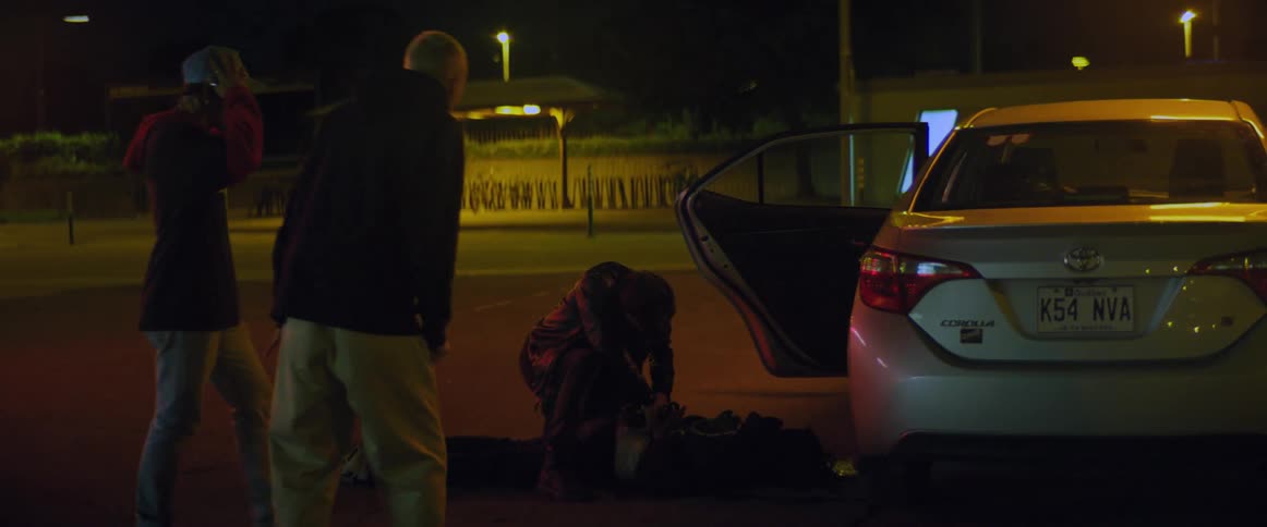 Two men wait their turn while another rifles pockets of person lying on ground near car with open door