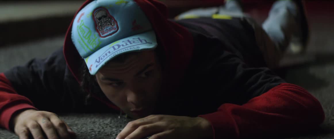 A teenage boy wearing a cap is lying on his stomach on a carpeted floor looking around in a sneaky manner