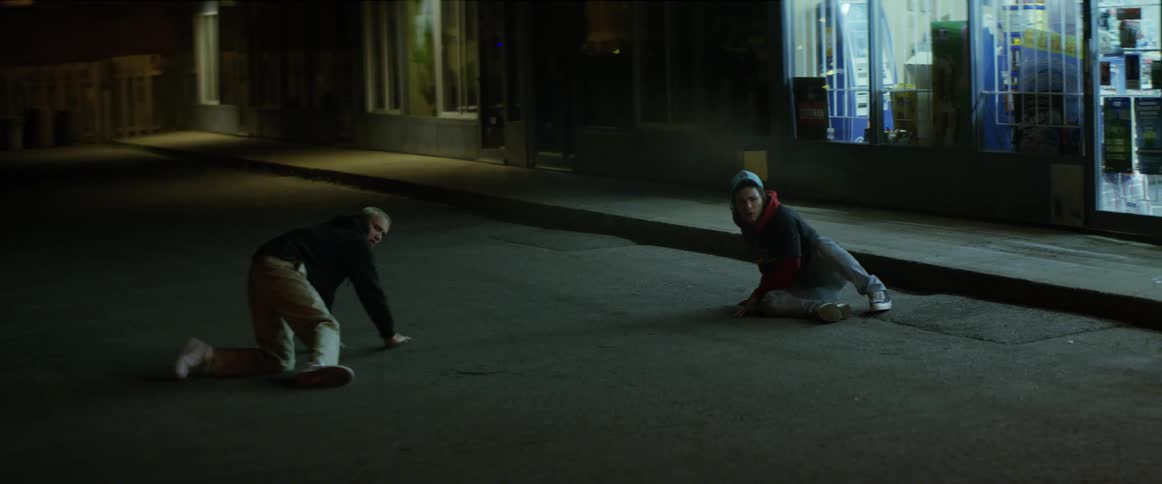 Two teenage boys lying on a street outside a store get up a to help a man struggling to get away from someone