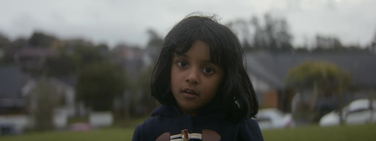 A child looks into the camera while standing outdoors in a residential neighborhood during the day
