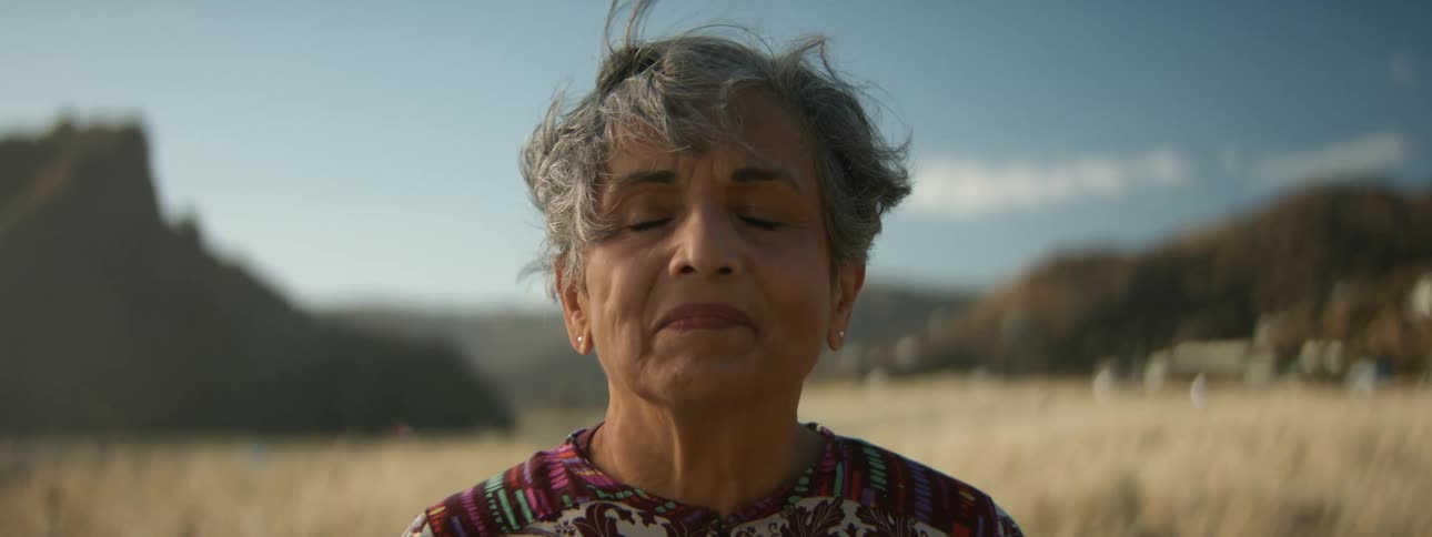 A woman has her eyes closed as she meditates in a wheat field as her hair moves in the breeze and there are mountains in the distance