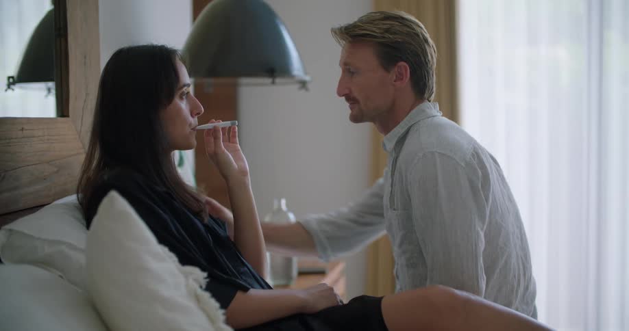 A man sits with a woman who is in bed while using a thermometer to take her temperature