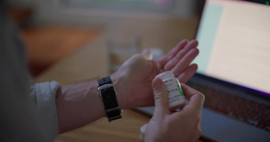 A man is sitting at a desk in front of a laptop computer and he opens a pill bottle and takes a pill then drinks from a glass of water