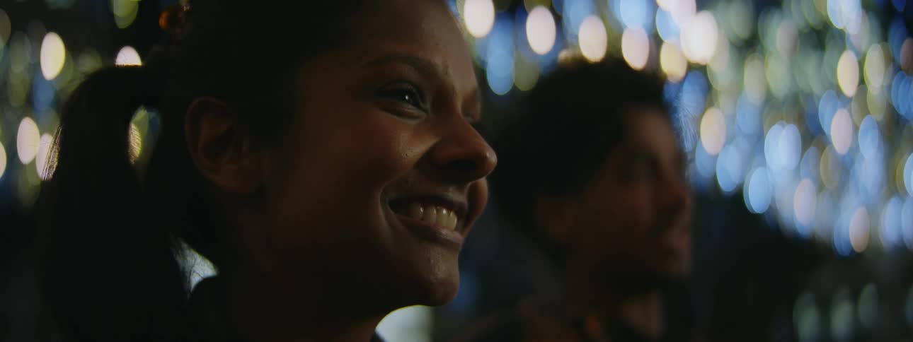 Two people stand smiling near tree strung with lights as people pass in the background