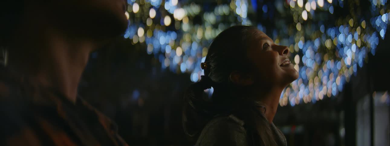 A teenage girl and boy are sitting on a bench and she is smiling as she looks up at the sky and there are bright lights behind them