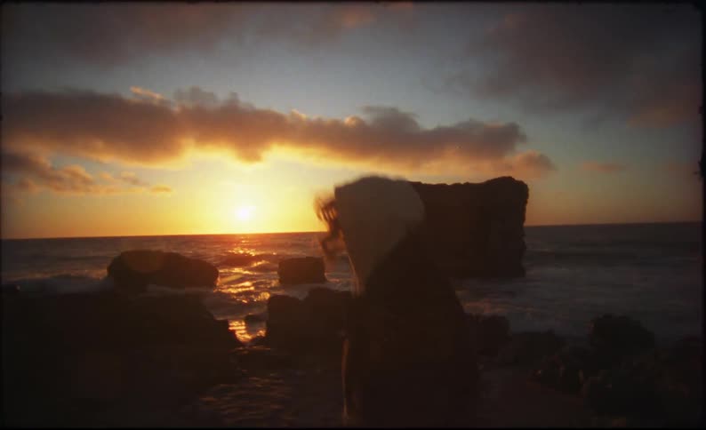Two people are walking on a beach as water splashes against rocks and the sun shines brightly on the horizon