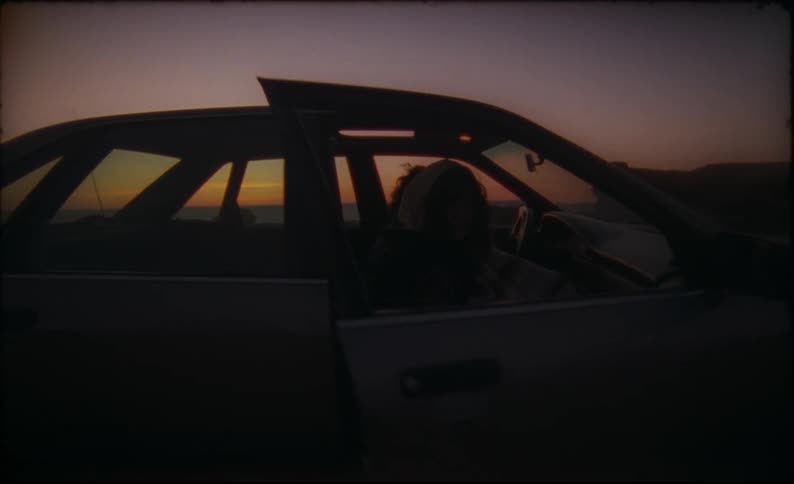 Two people parked in a car on a beach cover up with a blanket and lie their seats back to sleep