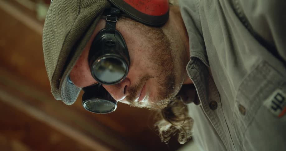 A man wearing dark goggles and earmuffs is standing in a room looking down
