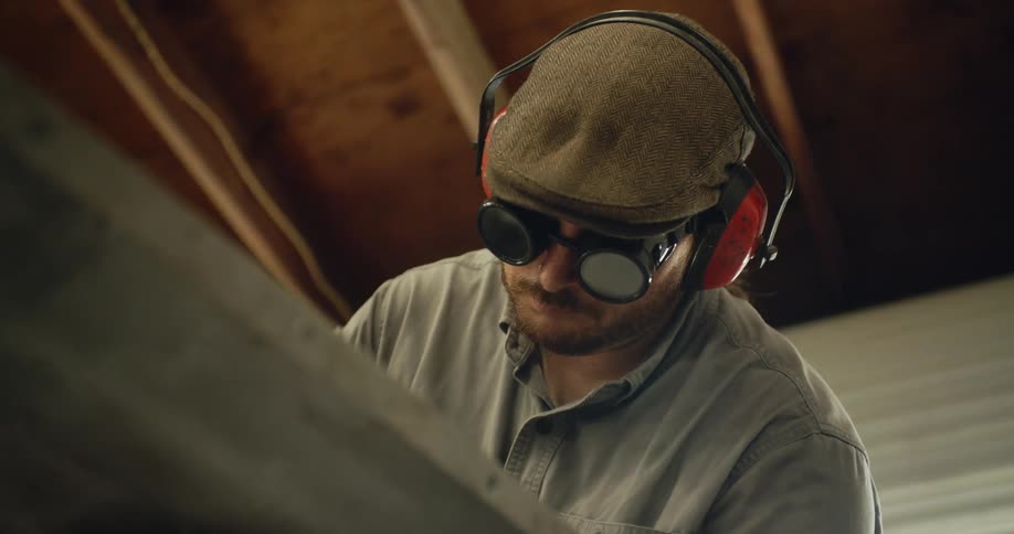 A man wearing dark goggles and earmuffs is standing in a room looking down at material he is working on