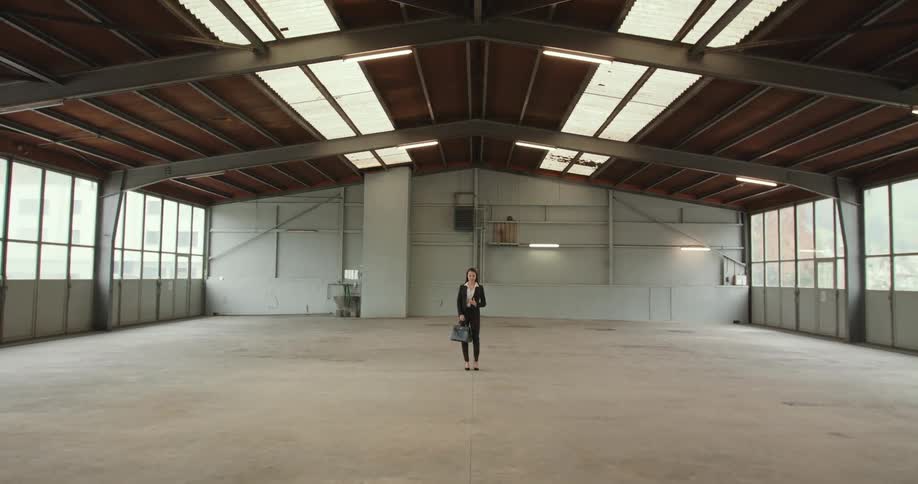 A woman wearing a black pant suit and holding a briefcase is standing in a large empty room