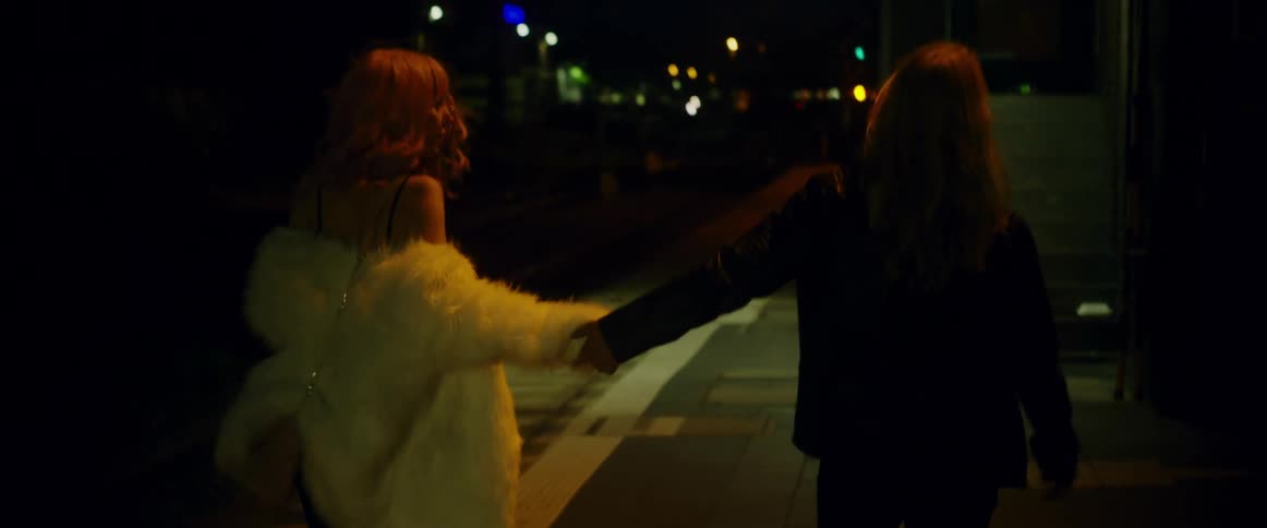 A redheaded woman wearing a white fur coat and carrying a wine bottle is walking near train tracks with a long haired man