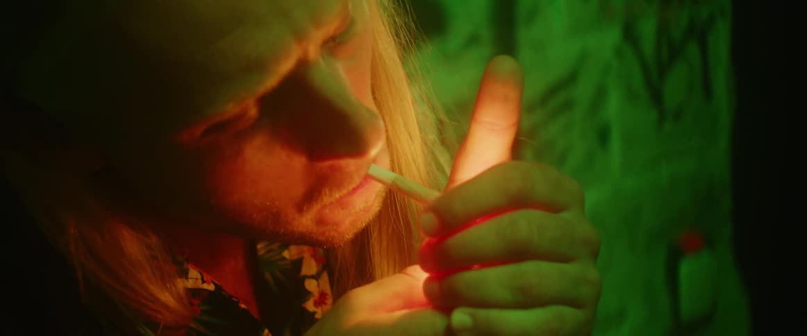 A man is leaning against a wall lighting a cigarette and smoking and there is a green light shining on him
