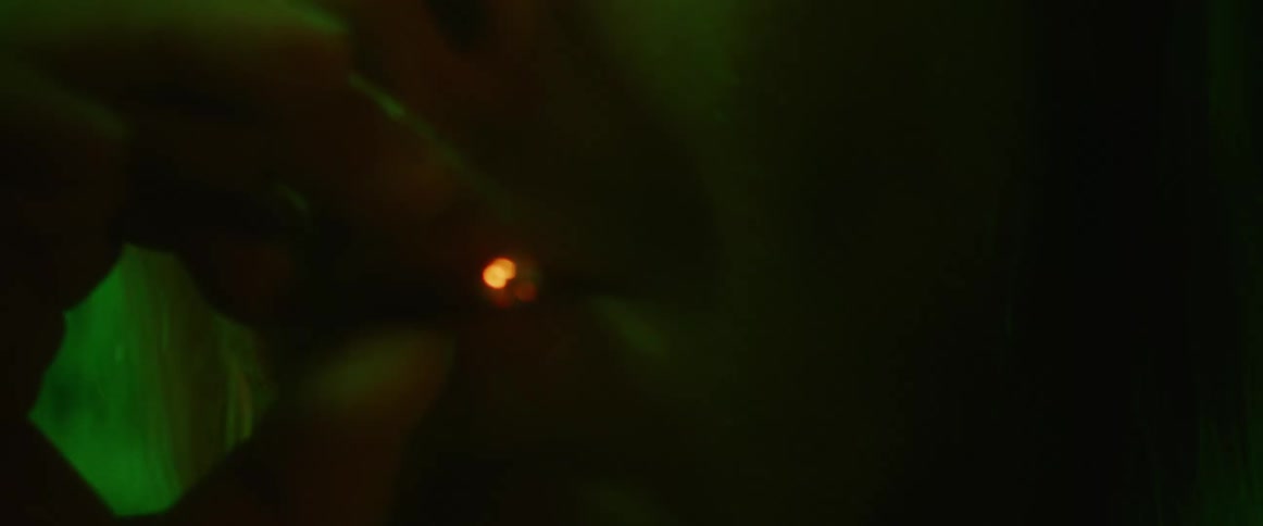 A man is leaning against a brick wall smoking and there is a green light shining on him