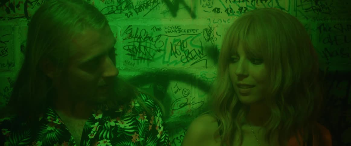 Man smokes as he and woman lean against graffiti covered wall in shadowy room lit by green lighting