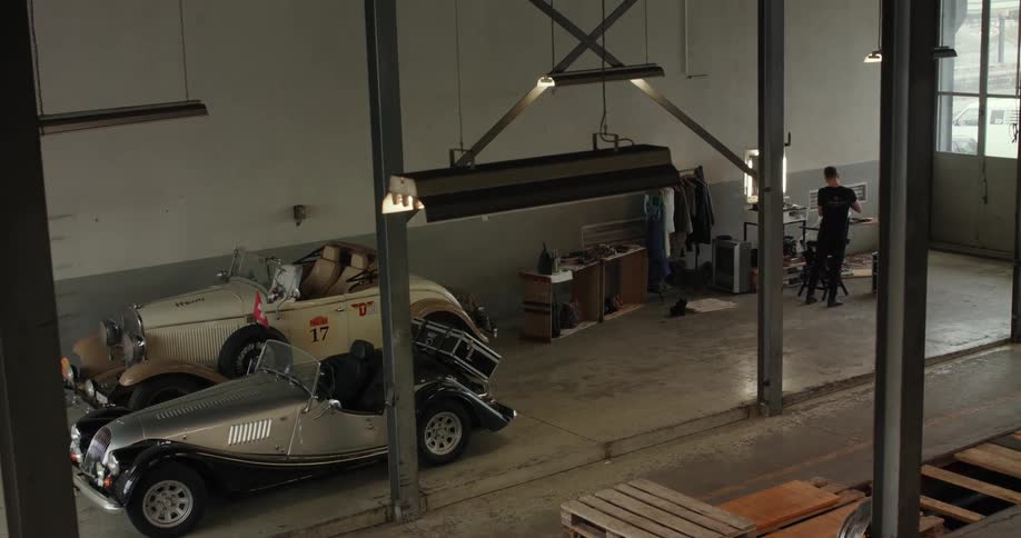 A person stands while working on a part in a garage with two vintage automobiles in it