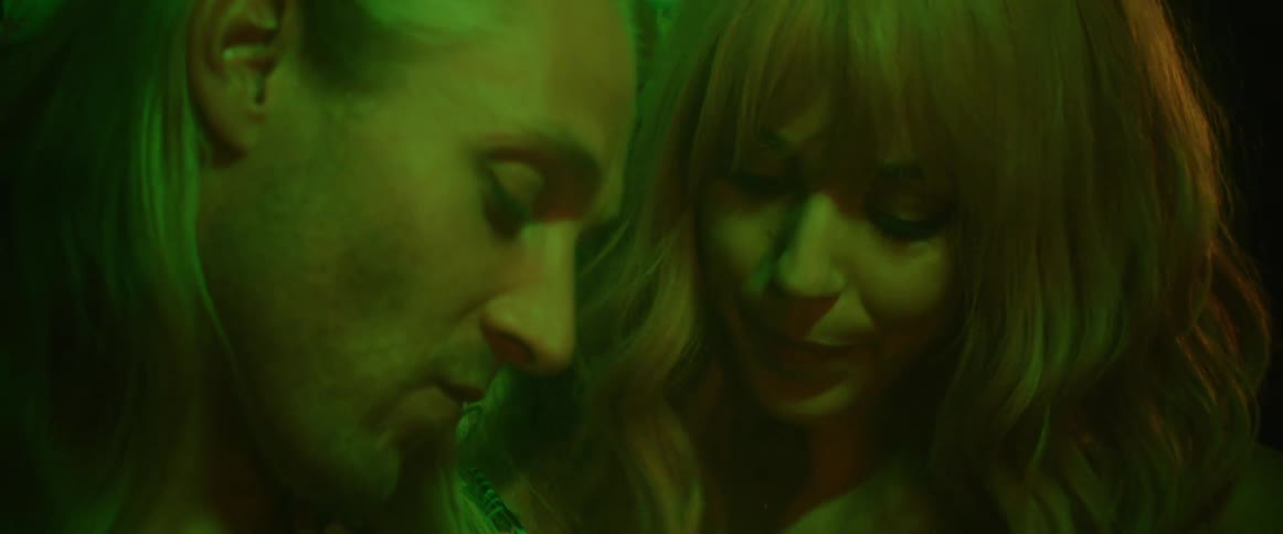 A man and a woman are sitting outside leaning against a building and she whispers into his ear and looks at him affectionately