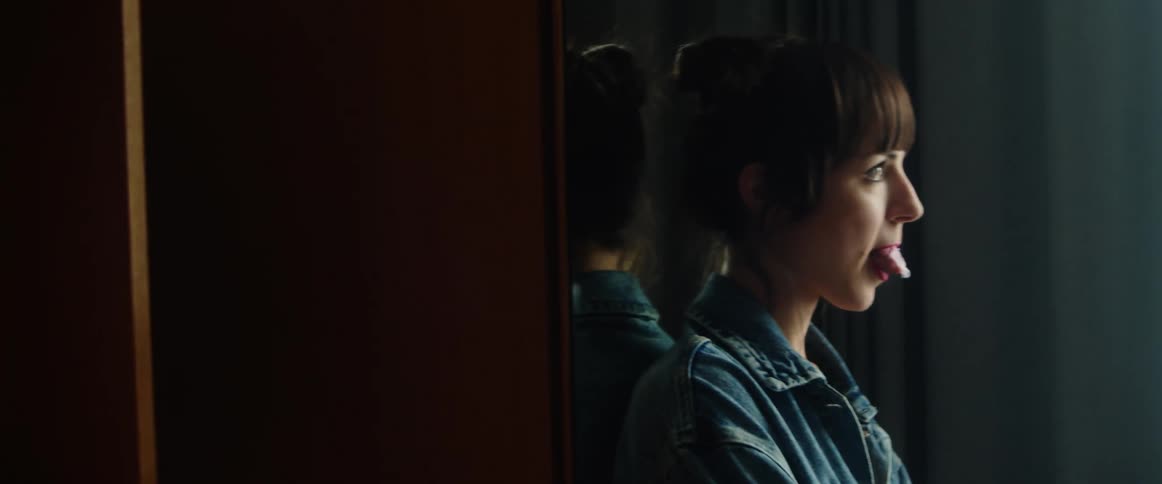 A teenage girl is standing in a room leaning against a mirror chewing bubble gum and blowing bubbles as she looks out the window