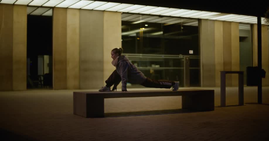 A woman is stretching and warming up on a bench in front of a building at night