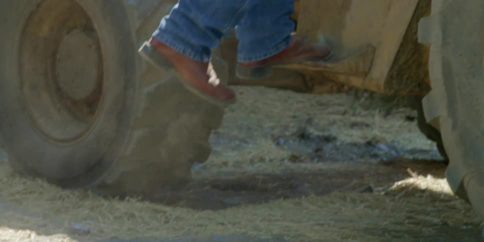 A man wearing boots walks through hay and enters a tractor