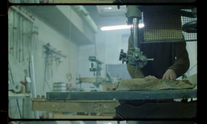 A man is in a workshop operating a saw on a table as he pushes a piece of wood and cuts it