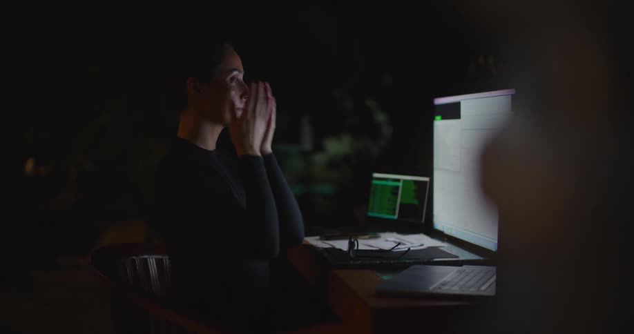 A woman takes off her places and places her hands on her face as she works at her computer at night