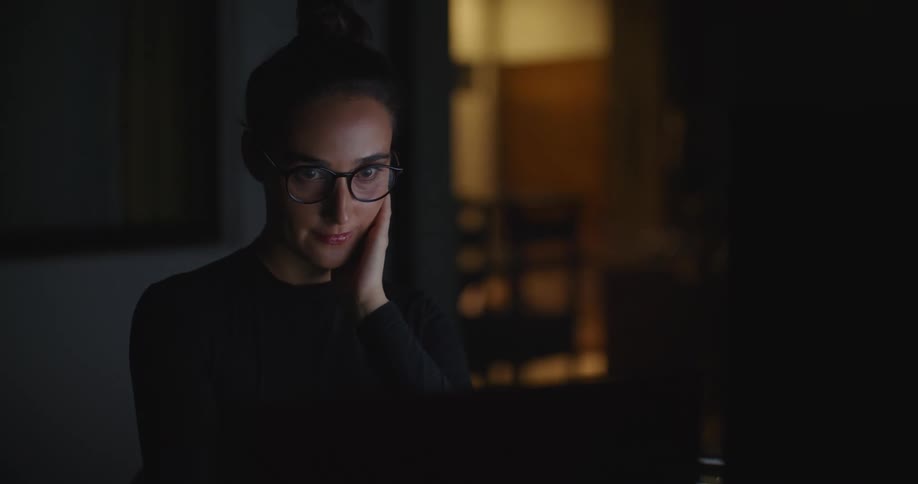 Woman concentrates on laptop screen in dark room as light shines from another room in the background