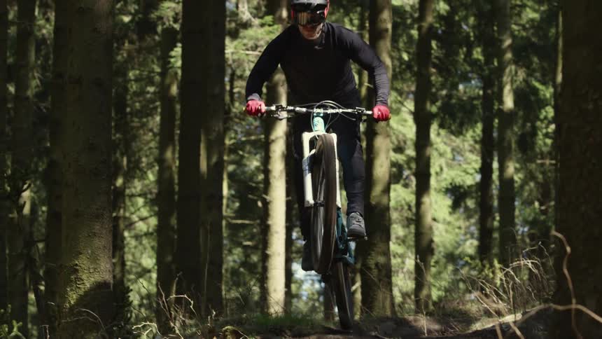 A cyclist repeatedly goes airborne while riding over an uneven dirt path through a woodland during the day