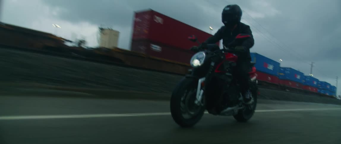 A cyclist dressed in black and wearing a black helmet is riding a motorcycle on a road alongside a train carrying cargo containers