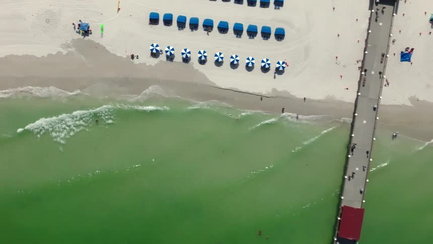 Tourists walk down a pier as others sunbathe on a sunny Florida beach, drone shot