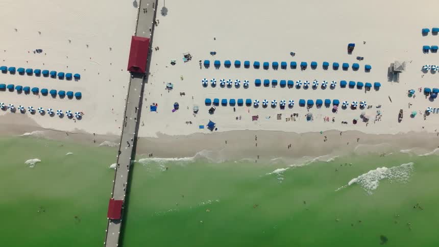 Beachgoers enjoy a sunny day on a Florida beach in the summer time, next to a long pier, drone shot