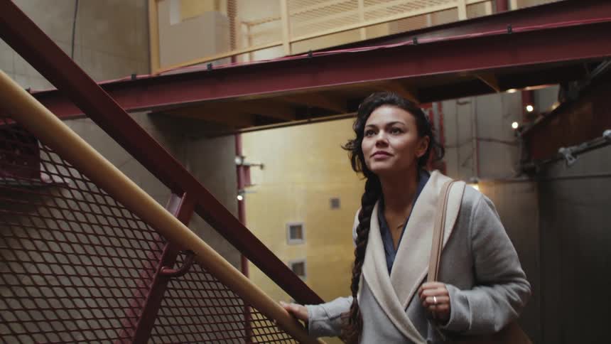 Woman wearing coat climbs stairs with a slight smile holding onto banister and plants can be seen in background on upper floor