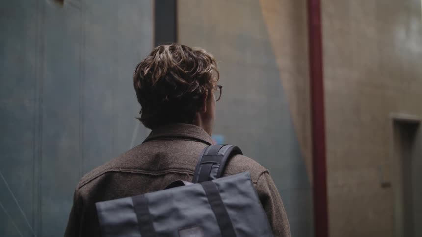 Man with wavy hair glasses brown jacketand backpack walks down the street looking up at apartments