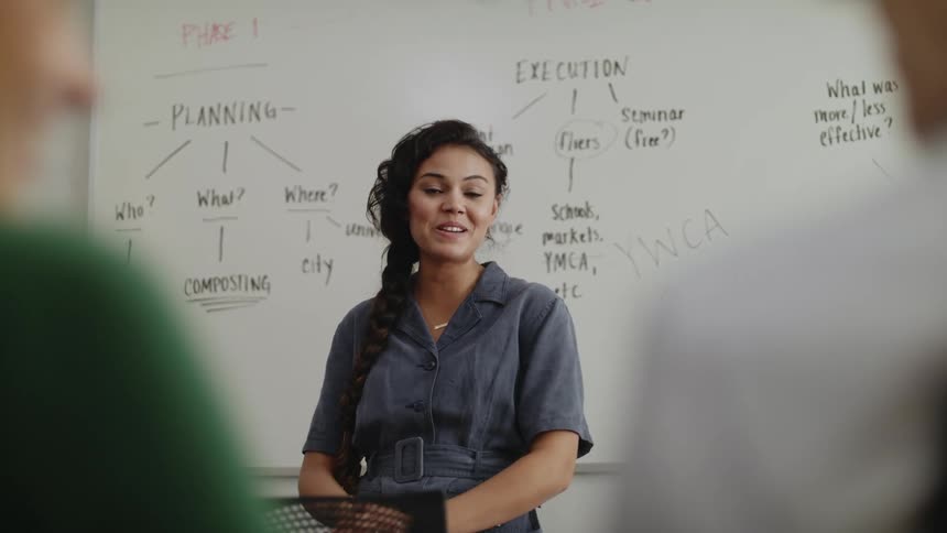 A woman in front of a class with people in the foreground with a whiteboard and writing in the background