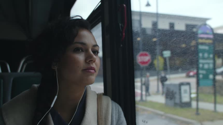 A woman with headphones in her ears sits on a bus and looks out the window as it drives past people and cars and buildings