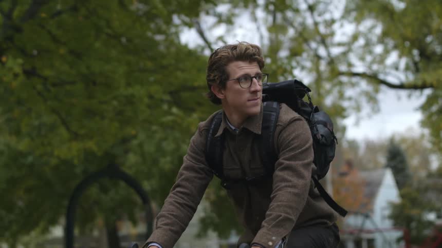 Man wearing jacket and backpack rides red bicycle along side of road through neighborhood