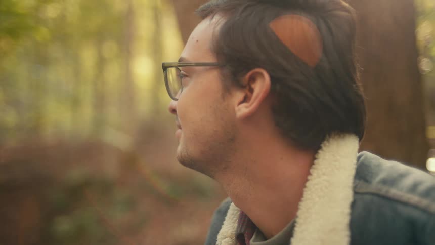 A young white man is sitting down around the forest area