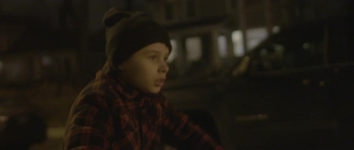 A fearful looking boy bicycles down a street at night