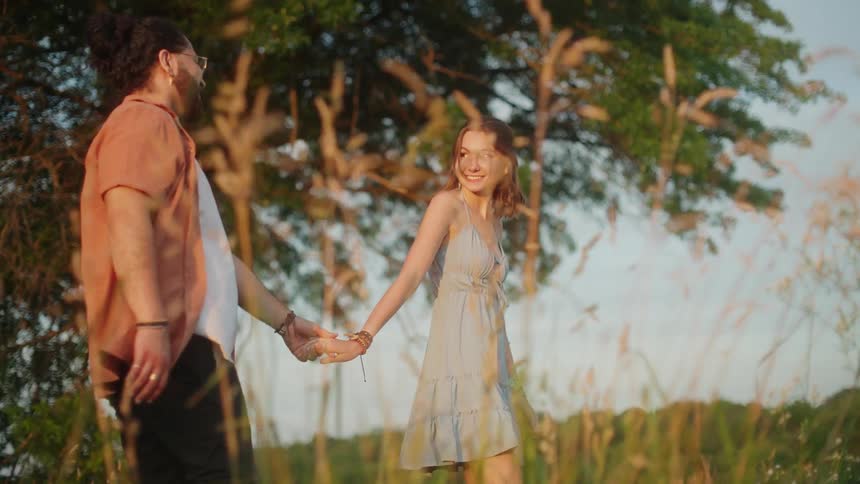 A woman smiles at her boyfriend while leading him by the hand through a field