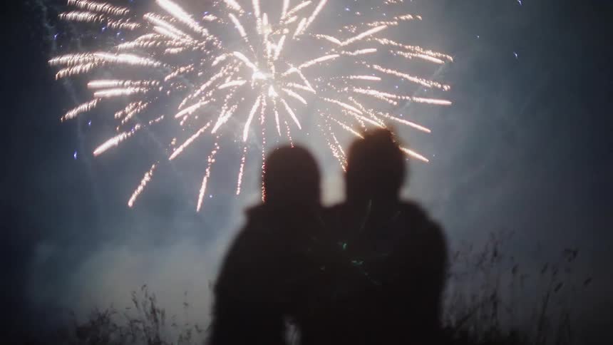 The silhouette of a couple sitting close in the grass watching fireworks explode in the night sky