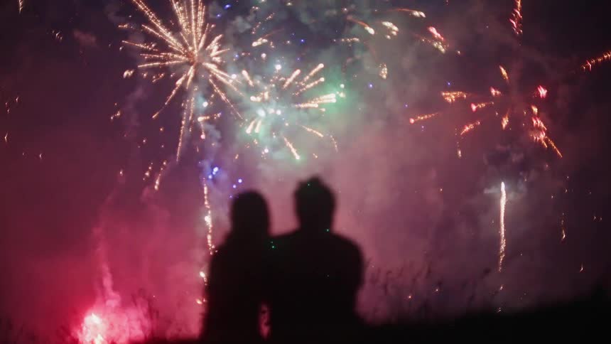A man and woman sit in a field together and watch fireworks light up the sky