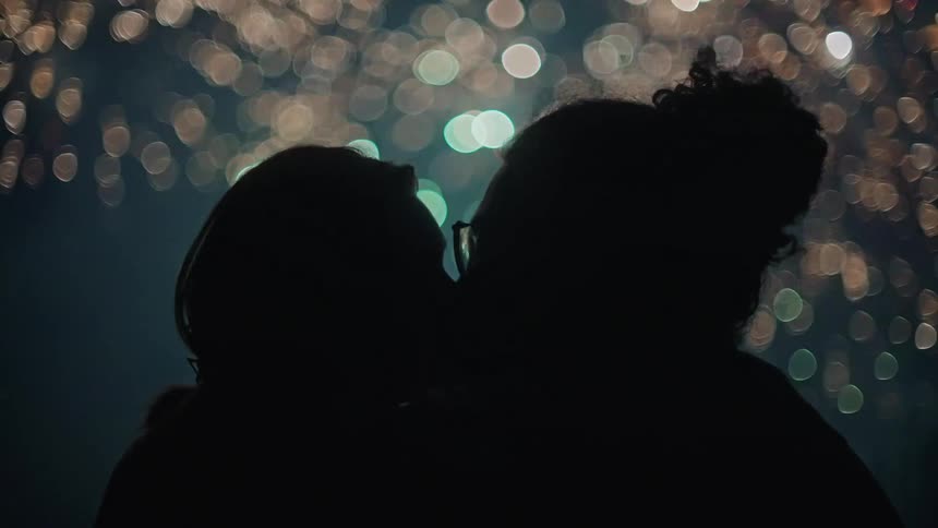 A man and a woman are outside kissing as bright and colorful fireworks fill the sky