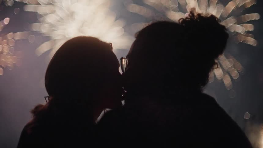A man and a woman are sitting outside kissing as bright and colorful fireworks fill the sky