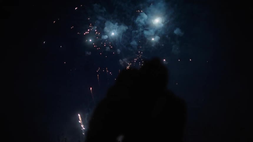 Silhouettes of a couple sitting together in the grass watching bright and colorful fireworks